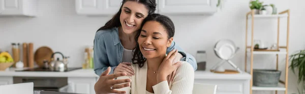 Happy interracial lesbian couple hugging each other in kitchen, banner — Stock Photo