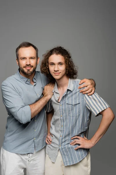 Feliz padre abrazando hombros de rizado adolescente hijo aislado en gris - foto de stock