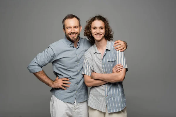 Happy bearded father hugging shoulder of positive teenage son isolated on grey — Stock Photo