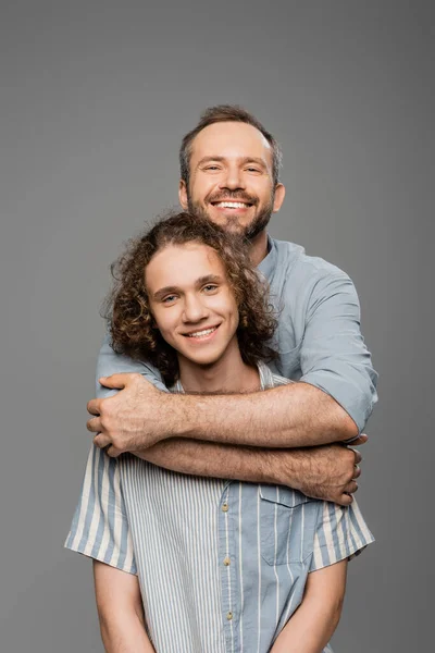 Cheerful man hugging curly teenager son and looking at camera isolated on grey — Stock Photo