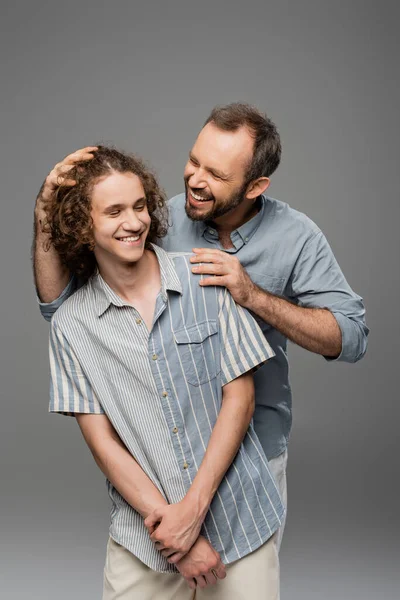 Alegre papá tocando pelo rizado de su hijo adolescente y sonriendo aislado en gris - foto de stock