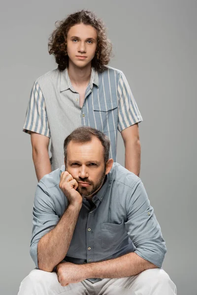 Bored father looking at camera next to curly teenage son isolated on grey — Stock Photo