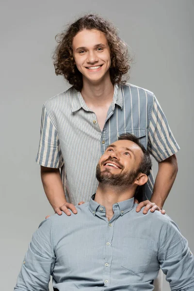 Positive father smiling and looking at happy teenage son isolated on grey — Stock Photo
