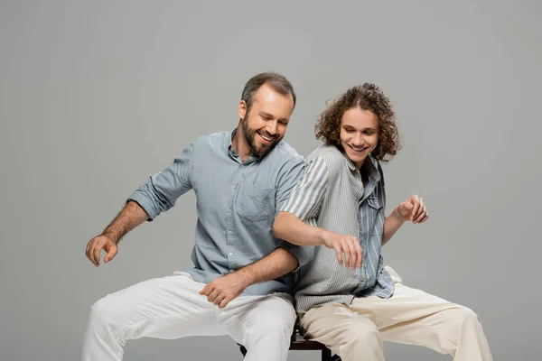 Щасливий батько і син розважаються і штовхають один одного, сидячи на одному стільці ізольовані на сірому — стокове фото