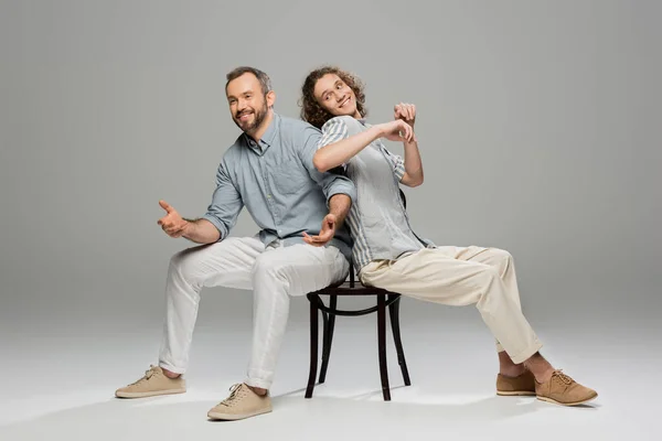 Happy father and carefree teenage son having fun and pushing each other while sitting on same chair on grey — Stock Photo