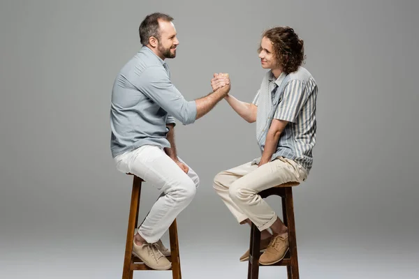 Vue latérale du père heureux et du fils adolescent serrant la main assis sur des chaises hautes sur gris — Photo de stock