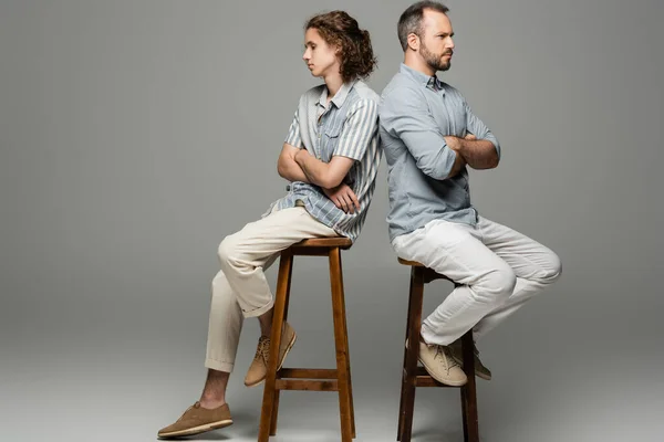 Обиженные отец и сын-подросток сидят со сложенными руками спина к спине на серой — стоковое фото