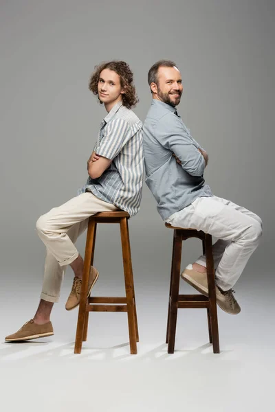 Fröhlicher Vater und Teenager-Sohn sitzen mit verschränkten Armen Rücken an Rücken auf grau — Stockfoto
