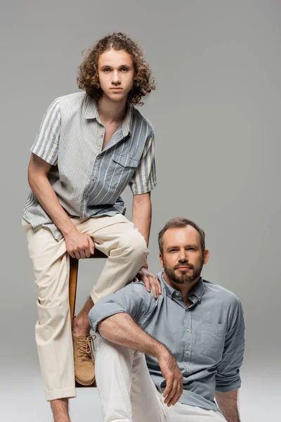 Curly teenage guy sitting on high chair next to bearded dad isolated on grey — Stock Photo