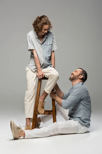 Rizado adolescente chico sonriendo mientras sentado en la silla alta al lado de feliz papá en gris - foto de stock