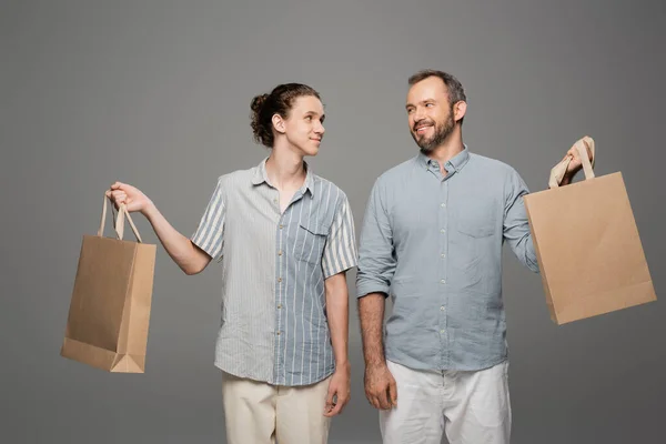 Happy teenage boy and cheerful dad holding shopping bags isolated on grey — Stock Photo