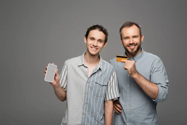 Щасливий батько тримає кредитну картку поруч з сином-підлітком з смартфоном в руці ізольовано на сірому — стокове фото