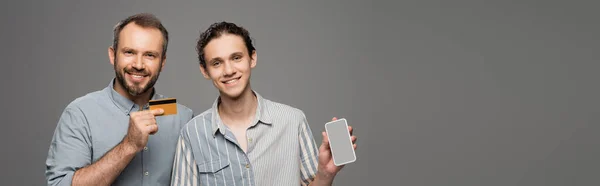 Glücklicher Vater mit Kreditkarte neben pubertierendem Sohn mit Smartphone in der Hand auf grauem Banner — Stockfoto