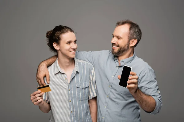 Glücklicher Vater mit Smartphone mit leerem Bildschirm neben pubertierendem Sohn mit Kreditkarte in der Hand isoliert auf grau — Stockfoto