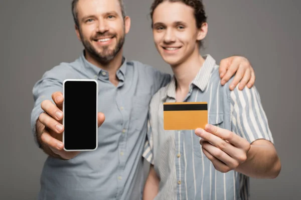 Счастливый отец держит смартфон с чистым экраном рядом с счастливым сыном-подростком с кредитной картой в руке, изолированной на сером — стоковое фото