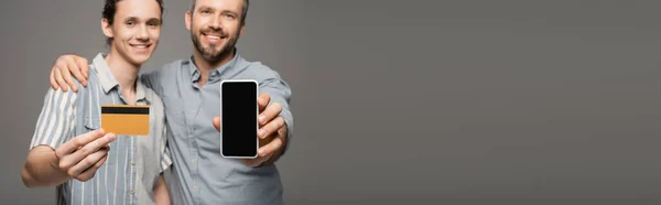 Heureux père tenant smartphone avec écran blanc à côté de heureux fils adolescent avec carte de crédit à la main isolé sur gris, bannière — Photo de stock
