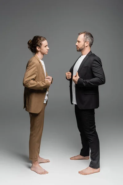 Longitud completa de padre e hijo bien vestidos ajustando las chaquetas y mirándose el uno al otro en gris - foto de stock
