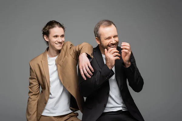 Cheerful teenage boy and overjoyed dad in suits laughing isolated on grey — Stock Photo