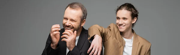Cheerful teenage boy and overjoyed dad in suits laughing isolated on grey, banner — Stock Photo