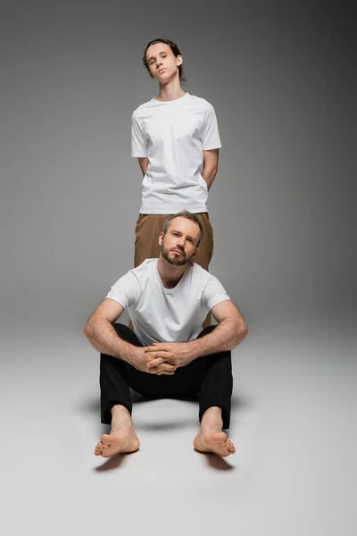 Pleine longueur de père barbu et fils adolescent en t-shirts blancs posant sur gris — Photo de stock