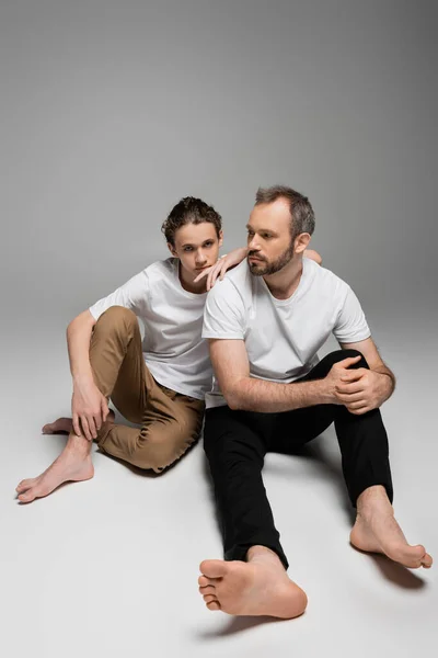 Longitud completa de padre descalzo e hijo adolescente en camisetas blancas posando sobre gris - foto de stock