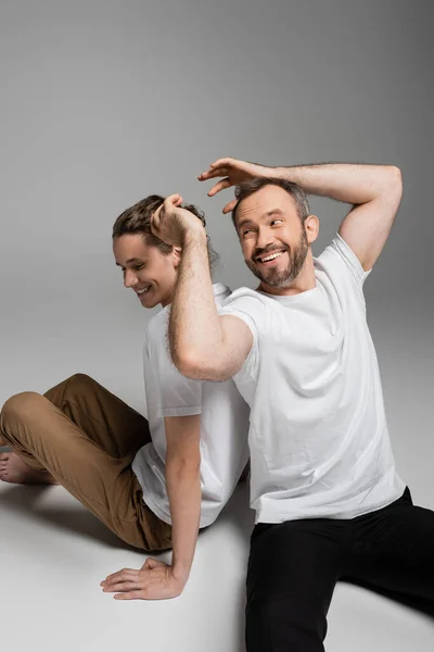 Cheerful father having fun with teenage son in white t-shirt and sitting on grey — Stock Photo