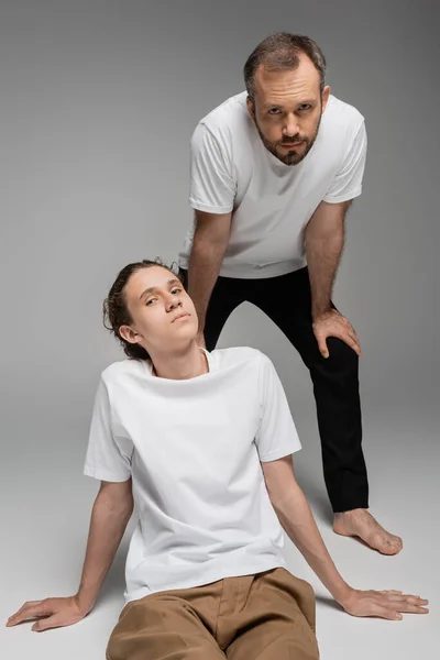 Бородатий батько і підліток син у білих футболках позує, дивлячись на камеру на сірому — стокове фото