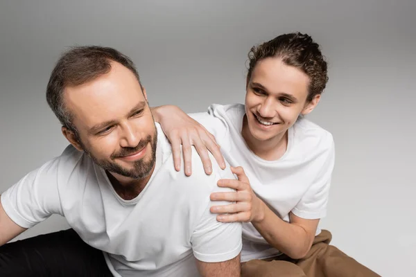 Lächelnder Vater und glücklicher Teenager-Sohn in weißen T-Shirts, die auf grau lächeln — Stockfoto