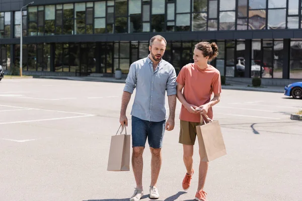 Pai barbudo andando ao lado de filho adolescente feliz e segurando sacos de compras perto do shopping — Fotografia de Stock