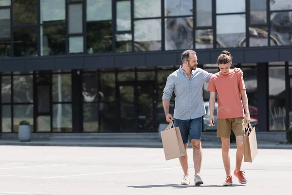 Glücklicher bärtiger Vater umarmt Teenager-Sohn, während er mit Einkaufstüten aus Einkaufszentrum läuft — Stockfoto
