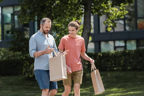 Hombre mirando dentro de la bolsa de compras cerca de feliz hijo adolescente al aire libre - foto de stock