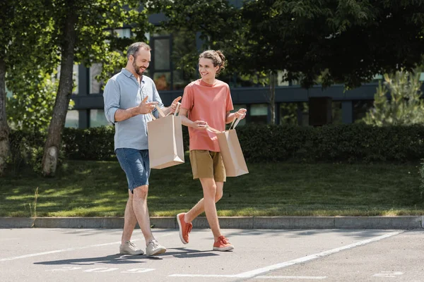 Pai feliz olhando para dentro do saco de compras enquanto caminha com o filho adolescente feliz ao ar livre — Fotografia de Stock