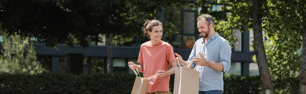 Padre feliz mirando dentro de la bolsa de compras mientras camina con el hijo adolescente feliz al aire libre, pancarta - foto de stock