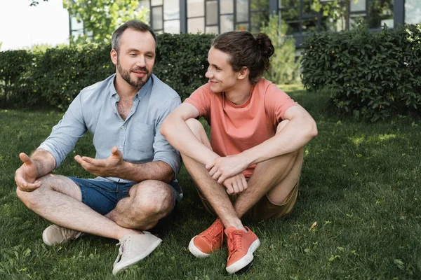 Happy bearded father and teenage son sitting together on green lawn — Stock Photo