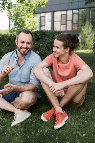 Cheerful bearded father and teenage son sitting together on green lawn — Stock Photo