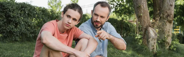 Bearded man pointing away near teenager son while sitting together in park, banner — Stock Photo