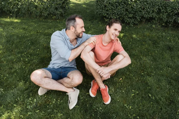 Blick von oben auf den glücklichen bärtigen Vater, der seinen Teenager-Sohn umarmt, während er zusammen auf dem grünen Rasen sitzt — Stockfoto