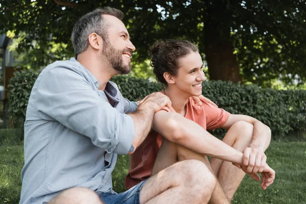 Щасливий бородатий батько обіймає усміхненого сина-підлітка і сидить разом на зеленому газоні — стокове фото