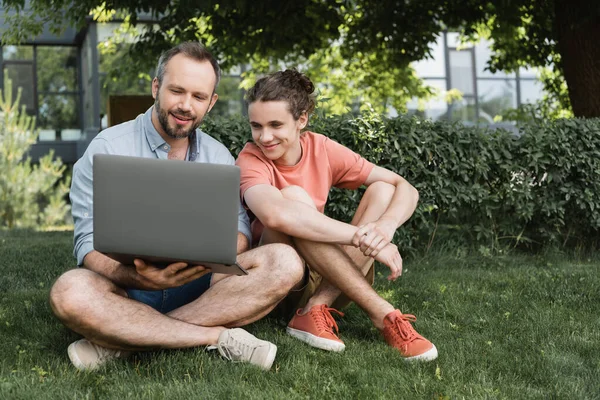 Padre feliz usando el ordenador portátil al lado del adolescente mientras están sentados juntos en el césped verde - foto de stock
