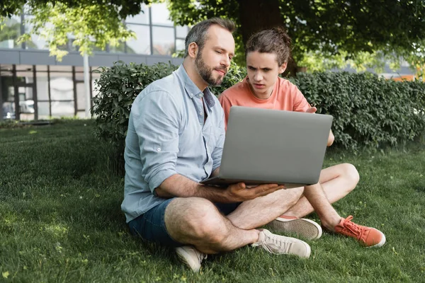 Батько і підліток син дивиться на ноутбук, сидячи разом на зеленому газоні — стокове фото
