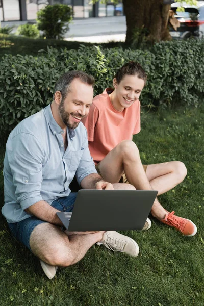 Pai feliz usando laptop ao lado de menino adolescente alegre enquanto sentados juntos no gramado verde — Fotografia de Stock