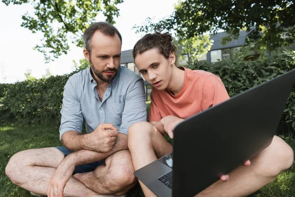 Vater und Sohn schauen auf Laptop, während sie zusammen auf grünem Rasen sitzen — Stockfoto