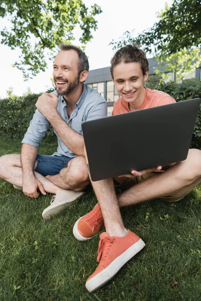 Feliz adolescente usando el ordenador portátil mientras está sentado junto con el padre en el césped verde - foto de stock