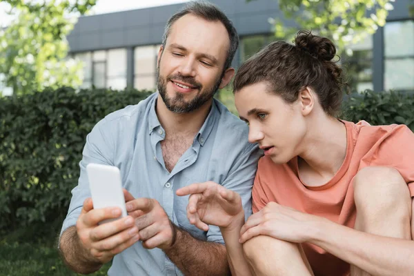 Adolescente apuntando con el dedo en el teléfono inteligente junto a padre feliz en el parque verde - foto de stock