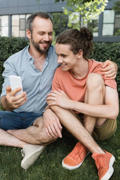 Щасливий чоловік тримає смартфон біля сина-підлітка, сидячи разом у зеленому парку — стокове фото