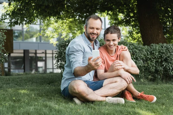 Счастливый мужчина делает селфи с сыном-подростком, сидя вместе в зеленом парке — стоковое фото