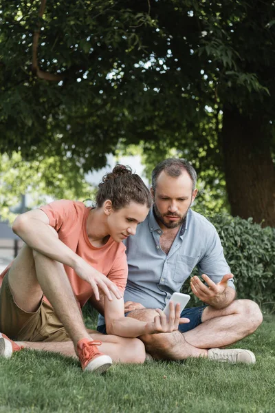 Отец и сын смотрят на смартфон, сидя вместе в зеленом парке — стоковое фото