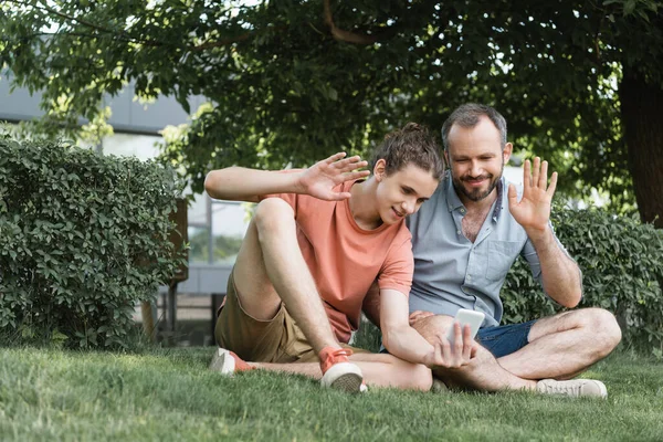 Pai feliz e filho adolescente acenando as mãos durante a chamada de vídeo enquanto sentados juntos no parque verde — Fotografia de Stock