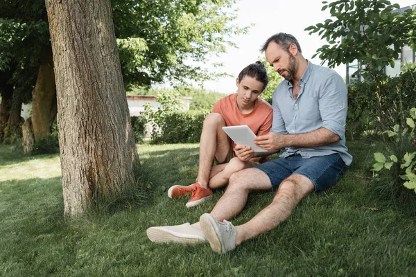 Barbudo padre e hijo adolescente mirando tableta digital mientras está sentado cerca del árbol en el parque - foto de stock
