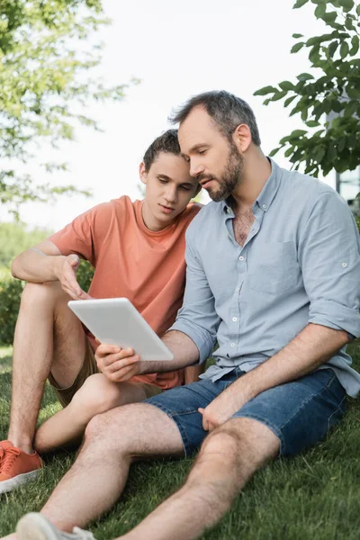 Бородатый отец и сын-подросток смотрят на цифровой планшет, сидя в зеленом парке — стоковое фото
